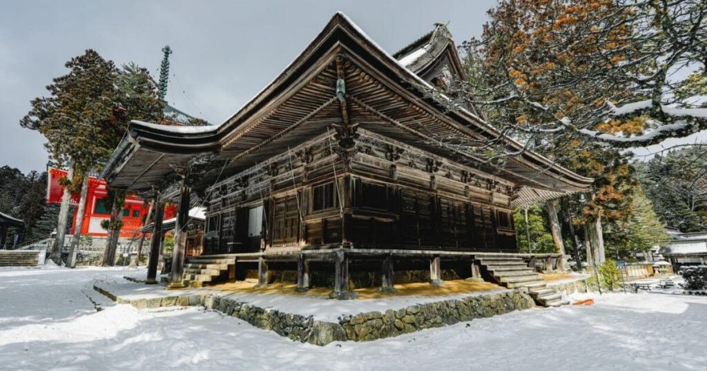 被白雪覆蓋的高野山寺院全景，木造建築與周圍杉木林映襯冬日景色。