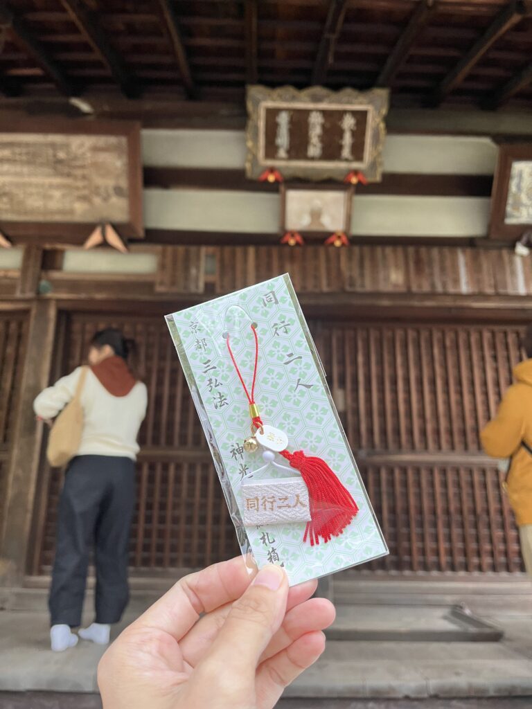 該圖片展示了在世界文化遺產京都神光院前，遊客手持精緻護身符的溫馨畫面。前景一隻手拿著裝在透明塑膠袋中的「納札箱」御守，御守吊卡上印有淡綠色幾何花紋、以及「同行二人」、「京都三弘法」與「納札箱」等綠色書法字樣。

御守主體為一條紅色吊繩，其上掛有一顆金色小鈴鐺、一片橢圓形白玉裝飾、一束鮮紅色的流蘇，以及一個寫有「同行二人」的白色迷你神札箱模型。背景為古色古香的神光寺，隱約可見斑駁的木質格柵門、上方懸掛著寫有弘法大師相關名號的深色木牌扁額，以及一位背著帆布包、正面向佛殿參拜的遊客背影，完美呈現出真言宗朝聖之旅的神聖與療癒感。