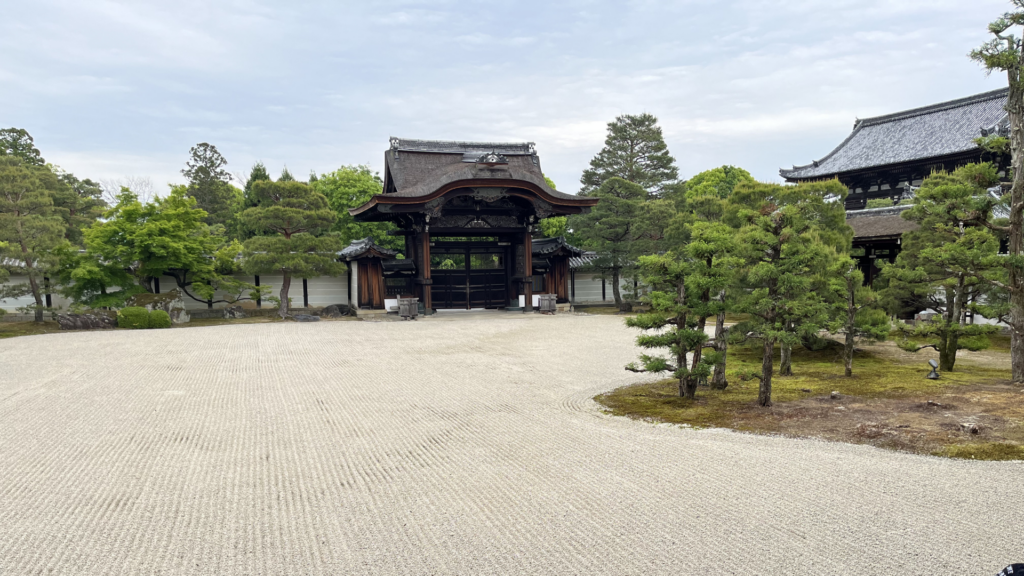 該圖片展示了世界文化遺產京都「仁和寺」御殿內氣勢高雅的「勅使門」與禪意枯山水庭園。畫面中央為一座極具代表性的傳統日式木造唐門（勅使門），擁有弧度優雅的檜皮葺屋頂與細緻的木雕裝飾，兩側連接著白牆與木造側門。

前景是一片極其寬闊、打理得井然有序的枯山水白砂地，表面耙制出細密整齊的平行砂紋；右側點綴著一片覆蓋著翠綠青苔、修剪圓潤的日式松樹與盆栽景觀。右後方隱約可見宏偉的二王門或金堂建築屋頂，天空呈現溫和的微陰微亮採光，整體構圖比例和諧，完美烘托出日本皇家御寺特有的莊嚴、寧靜與禪宗美學意境。