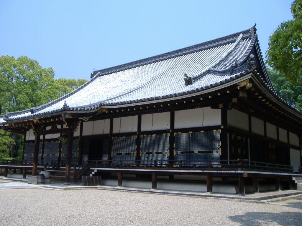 該圖片展示了京都世界文化遺產「仁和寺」被列為日本國寶的「金堂」側面全景。主體結構為一座自京都御所的紫宸殿移築而來的桃山時代宮殿風格木造建築，上覆精美緻密的黑灰色瓦片入母屋造屋頂（歇山頂），展现出古典高雅的對稱美學。殿身採用古樸的深色木質樑柱，牆面由白泥牆與精緻的格子窗櫺、帶有金箔裝飾的蔀戶（吊窗）交織而成，殿前設有帶有台階的優雅木造迴廊。 大門前方是一片開闊平坦的灰色碎石廣場，左側與右後方環繞著茂密翠綠的夏日林木。在澄澈明亮的蔚藍晴空烘托下，整幅畫面完美重現了這座與日本皇室擁有深厚淵源的國寶級佛殿所特有的肅穆、莊嚴與宮廷建築美學。