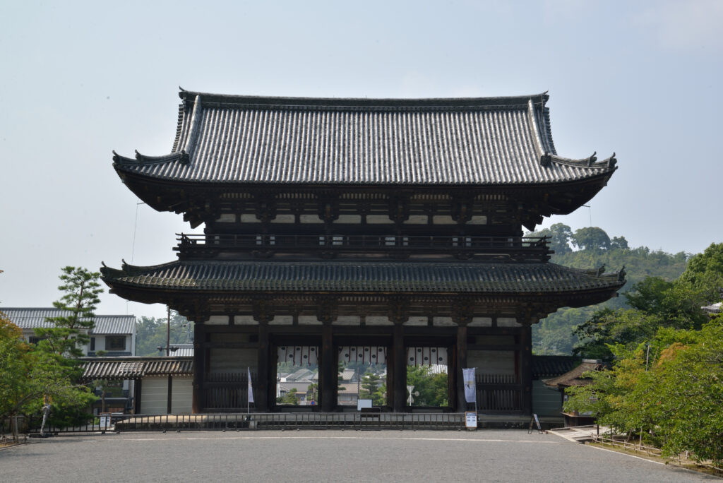 該圖片展示了京都世界文化遺產「仁和寺」氣勢磅礡的二王門（Nio-mon Gate）正面全景。主體結構為一座歷史悠久、規模宏大的兩層日式木造重簷歇山頂城門式建築，上覆整齊緻密的黑灰色瓦片屋頂，展現出純粹古樸的木質色澤。門下設有三個通道，內部掛著帶有紅白條紋的傳統帷幔。 大門前方是一片開闊、平坦的灰色碎石廣場，左側點綴著一株挺拔的松樹與白牆民居，右側則被茂密翠綠的林木所環繞，遠景隱約可見蔥鬱的群山天際線。在晴朗均衡的自然光線下，整幅畫面盡顯這座與知恩院三門、南禪寺三門並稱為「京都三大門」的江戶時代代表性建築特有的莊嚴、沉靜與古建築美學。