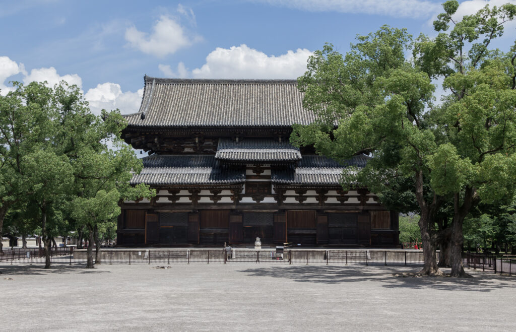 該圖片展示了位於日本京都的世界文化遺產東寺(教王護國寺)之主殿「金堂」全景。畫面中央為一座氣勢磅礡的雙層桃山時代風格木造大殿,建築擁有歷經歲月洗禮的古樸深色木質樑柱、精緻的白牆斗拱結構,以及宏偉的灰瓦歇山頂屋頂。大殿前方是一片寬闊平坦、打理整潔的灰色砂石廣場,四周環繞著低矮的金屬防護圍欄,兩側點綴著多棵茂密翠綠的百年老樹。在晴朗明亮、帶有幾朵白雲的藍天襯托下,充分展現出這座日本國寶級寺院歷史悠久的莊嚴與沉靜之美。