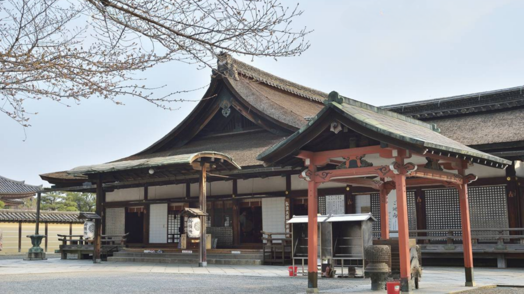 該圖片展示了位於京都的世界文化遺產「東寺」(教王護國寺)境內歷史悠久的御影堂(又稱大師堂)外觀景致。畫面中央的主體建築為一座極具寢殿造風格的古老木造大殿,擁有質樸獨特的檜皮葺屋頂,呈現出沉穩的棕褐色澤與優雅的屋脊線條。殿前左側設有懸掛著白色日式提燈的雨棚入口,右前方則矗立著一座漆有橙紅色澤的傳統木造供奉亭(香爐亭),亭下配有不鏽鋼置物架與祭祀器皿。